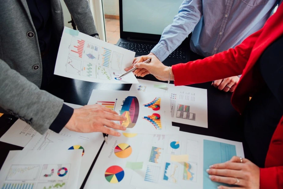 Colleagues reviewing printed business reports and colorful charts on a table during a collaborative data analysis meeting.