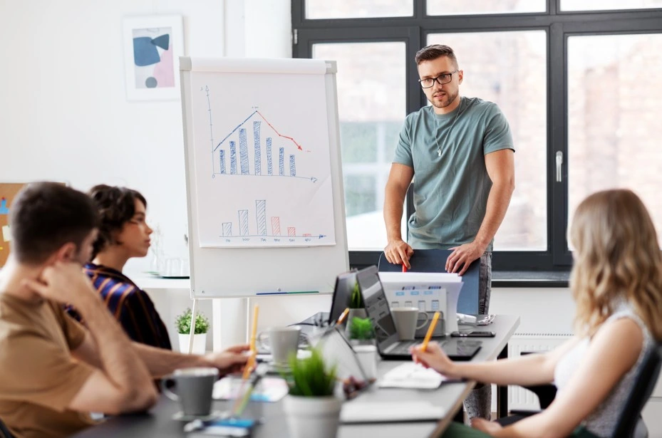 Team brainstorming session with presenter explaining bar and line charts on a flipchart during a business meeting.