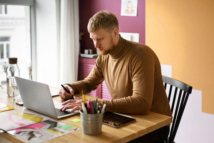 Focused man working on laptop with notes and office supplies at creative desk