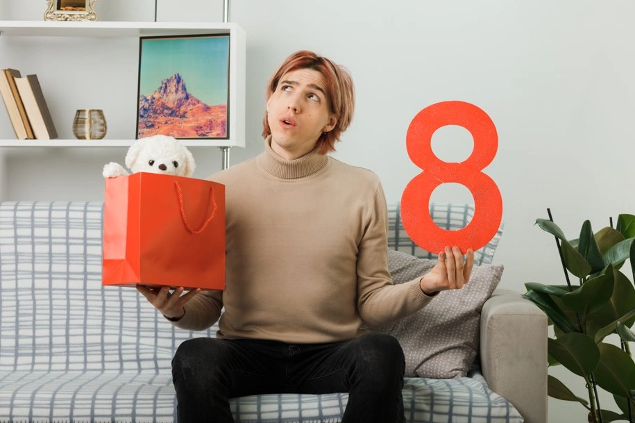 Young man comparing gift options while holding a large number eight sign in a modern living room.